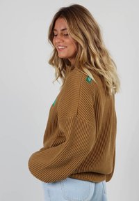 Pull marron côtelé avec une coupe décontractée et un col rond. Doté d'une petite étiquette verte à l'épaule. Associé à un jean bleu clair.