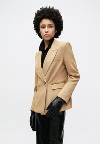 Μπεζ σακάκι με εγκοπές στο πέτο, διπλά κουμπιά και χρυσή αλυσίδα στη μέση. Φορεμένο πάνω από μαύρο τοπ και συνδυασμένο με μαύρα γάντια.