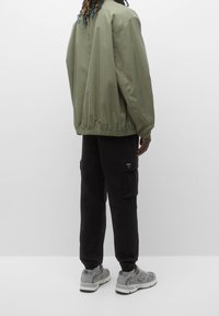 Olijfgroene bomberjack met elastische manchetten, gecombineerd met zwarte cargobroek en grijze sneakers. Gladde stof en een ontspannen pasvorm.