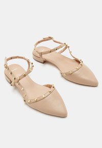 Bailarinas nude de punta afilada con acentos de tachuelas doradas, que presentan un diseño de tiras y un tacón bajo para mayor confort. Textura de cuero suave.