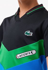 Black and green short-sleeve polo shirt with a V-neck, featuring a Lacoste logo and blue color-blocked mesh panel at the bottom.