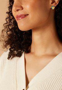 Goldene herzförmige Ohrstecker mit Türkisakzenten, die eine glatte Oberfläche und klare Schriftzüge aufweisen, am linken Ohr getragen, passend zu lockigem Haar.