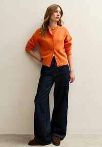 Pomarańczowy dziergany kardigan z okrągłym dekoltem i zapięciem na guziki, w połączeniu z szerokimi ciemnogranatowymi dżinsami i brązowymi loafersami, stojący na tle neutralnej ściany.
