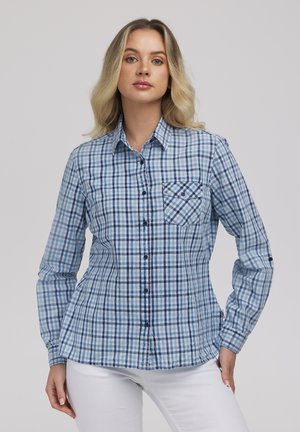 Kobieta ubrana w niebiesko-białą, kraciastą koszulę na długi rękaw z kieszenią na piersi, w połączeniu z białymi spodniami, na jednolitym tle.