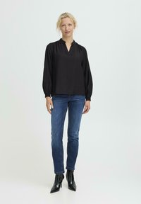 Schwarze Bluse mit langen Ärmeln, V-Ausschnitt und gerafften Akzenten, kombiniert mit dunkelblauen, figurbetonten Jeans und glänzenden schwarzen High-Heels.