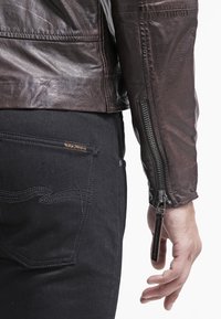 Giacca in pelle marrone con polsini con zip e finitura testurizzata, abbinata a jeans neri con un sottile motivo cucito sulle tasche.