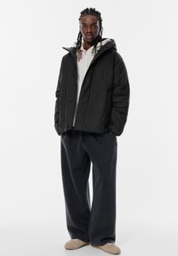Giacca a vento nera con cappuccio, dotata di chiusura a bottoni e vestibilità comoda. Abbinate a pantaloni larghi grigio scuro e scarpe beige.