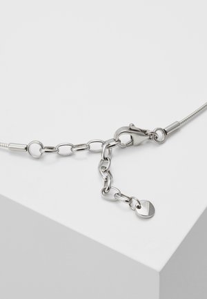 Fermoir de collier en chaîne argentée avec des maillons réglables et une petite plaque métallique ronde sur une surface blanche.