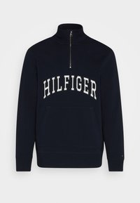 Sweat-shirt bleu marine à demi-fermeture éclair avec col montant, poche avant, et texte "HILFIGER" en grandes lettres blanches sur la poitrine.