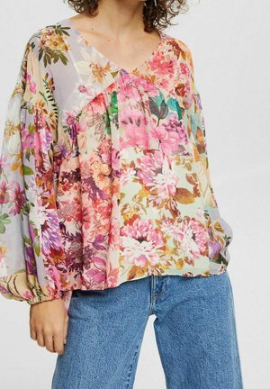 Vrouw die een losse blouse met lange mouwen draagt, bedrukt met meerkleurige bloemenpatronen, gecombineerd met blauwe spijkerbroeken.