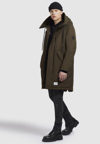 Parka verde închis cu glugă, buzunare frontale și șnururi. Purtați peste un pulover negru și blugi gri, asortați cu cizme negre.