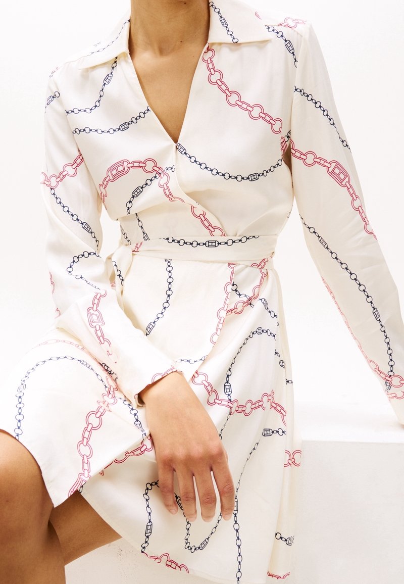 Femme portant une robe chemise blanche avec un motif chaîne rouge et noir, assise avec une main reposant sur son genou et ses genoux.