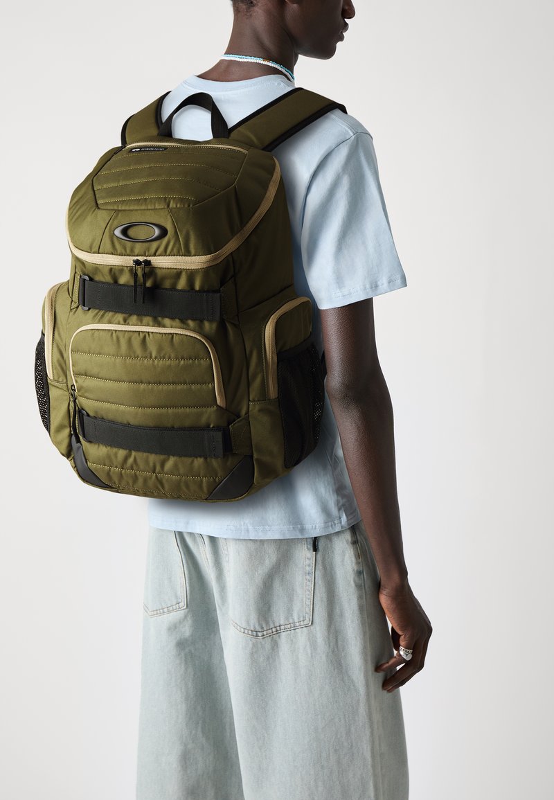 Zaino color verde oliva con sezioni imbottite, diverse tasche con zip, spallacci regolabili e tasche laterali in rete. Indossato su una camicia azzurro chiaro.