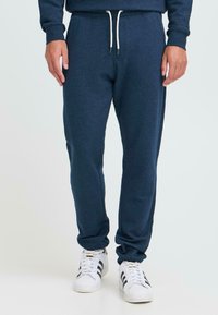 Hombre con pantalones de chándal azul marino y cordón blanco, y zapatillas blancas con franjas negras, de pie contra un fondo liso.