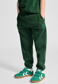 Grüne Jogginghosen aus weichem Stoff, mit einem Logo-Patch und seitlichen Taschen. Kombiniert mit grünen und weißen Klettverschluss-Sneakern.