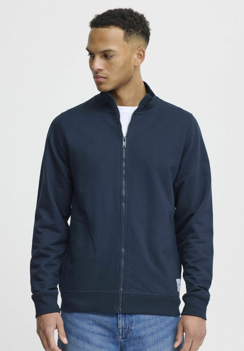 Sötétkék cipzáras pulóver, puha anyagból készült. Álló gallérral, két elülső zsebbel és bordás mandzsettával rendelkezik. Egyszerű, letisztult design.