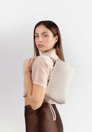Donna con capelli castani lunghi che indossa una blusa beige chiaro e una gonna marrone, porta una borsa beige sulla spalla, guardando oltre la spalla.