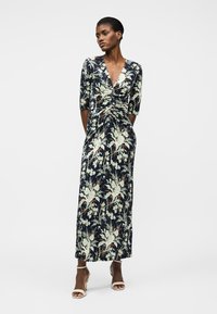 Γυναίκα όρθια φορώντας μακριά μπλε φόρεμα με λουλούδια, με ρυτιδωμένο εμπρόσθιο μέρος και μανίκια μέχρι τον αγκώνα, συνδυασμένο με μπεζ ψηλοτάκουνα πέδιλα με λουράκι στον αστράγαλο.