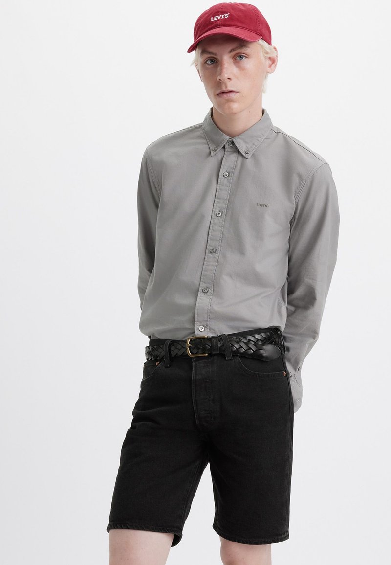 Camisa gris de botones, pantalones cortos de mezclilla negros, cinturón trenzado negro, gorra roja con el logo de "LEVI'S". La textura es suave, la tela es informal.