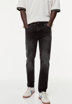 Homme portant un jean noir, une chemise blanche à manches courtes, des baskets blanches avec des bandes bleues, debout les mains dans les poches sur fond blanc.