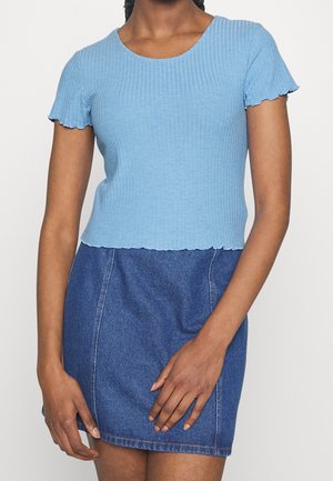Kvinde iført en lyseblå, ribbet kortærmet top og en mørkeblå denim nederdel, stående med hænderne afslappet foran.