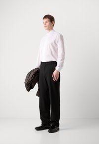 Fehér ing, plisszírozott elülső résszel, fekete elegáns nadrággal és fényes fekete cipővel; egy barna bőrdzsekit tart.