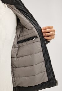 Schwarze Lederjacke mit einem grauen, gesteppten Futter und einer Reißverschlusstasche. Das Innere besteht aus glattem Stoff mit gepolstertem Material. Silberne Reißverschlussbeschläge.