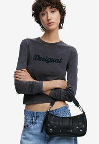 Borsa a tracolla in pelle nera con design testurizzato, borchie argentate e forma curvata. Indossata con pantaloni shorts in denim blu a vita alta e una maglietta grigia a maniche lunghe.