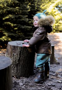 Brown waterproof coat with fur trim, light blue hat, and turquoise pants. Wearing black rubber boots. Standing beside a wooden stump.
