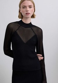 Mujer con cabello rubio corto, vestida con una blusa negra de canalé con mangas y canesú a rayas transparentes, posando contra un fondo claro y liso.