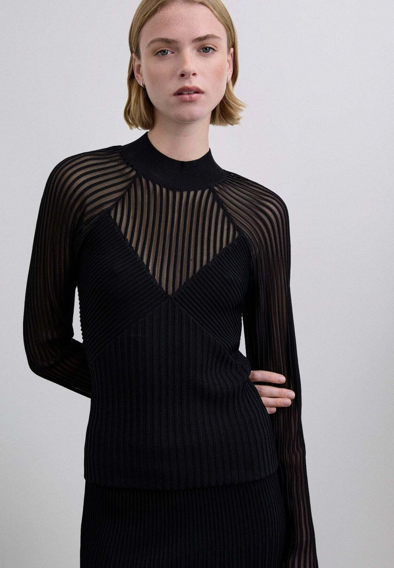 Mujer con cabello rubio corto, vestida con una blusa negra de canalé con mangas y canesú a rayas transparentes, posando contra un fondo claro y liso.