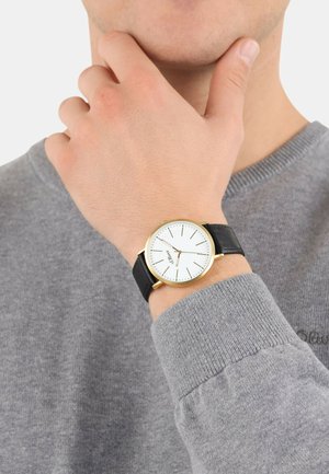 Goldfarbene Uhr mit einem weißen Zifferblatt und braunem Lederarmband, getragen an einem Handgelenk mit einem grauen Pullover, der das schlichte Design der Uhr hervorhebt.