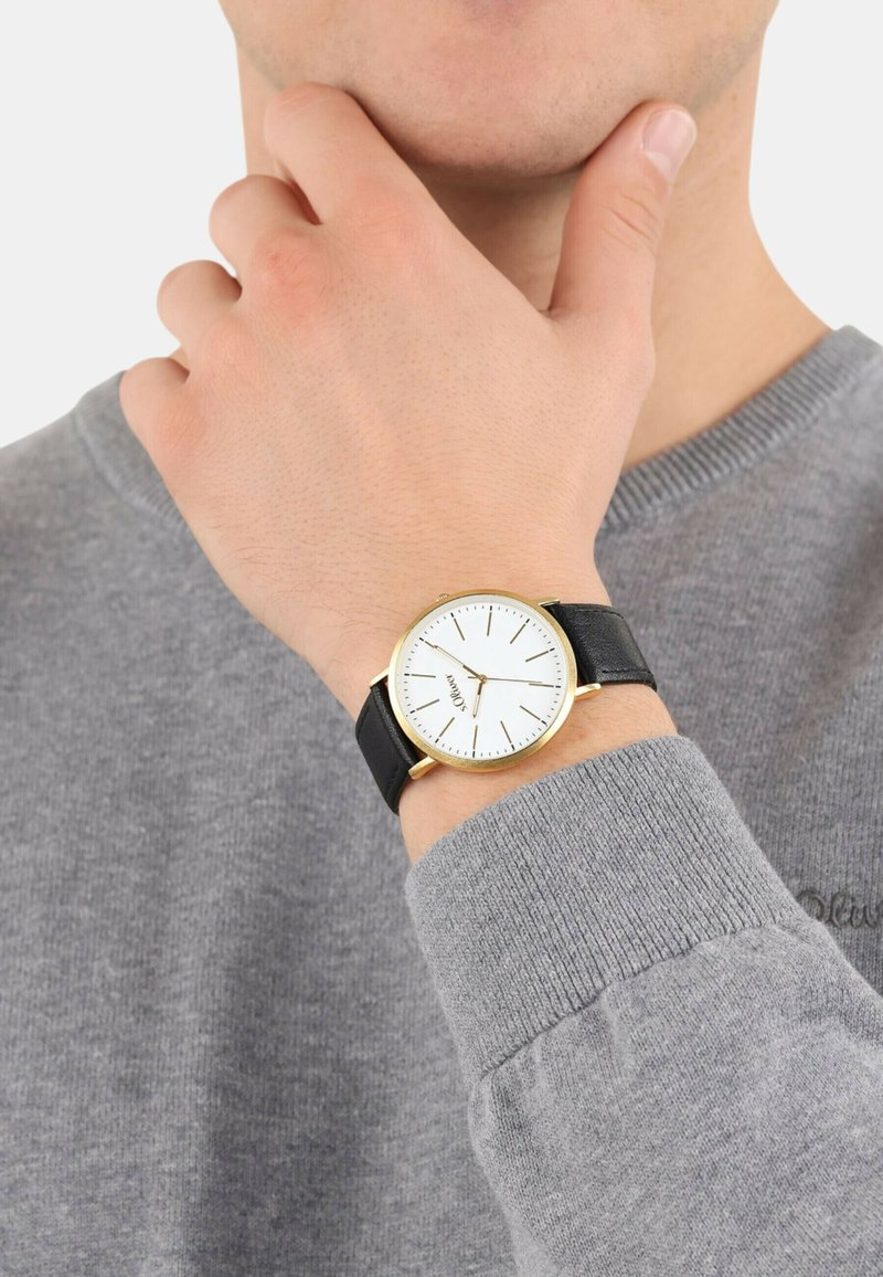 Gouden horloge met een witte wijzerplaat en zwarte leren band, gedragen om een pols met een grijze trui, waarbij het eenvoudige ontwerp van het horloge wordt benadrukt.