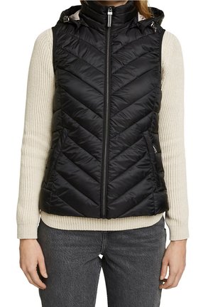 Musta tikattu hihaton toppaliivi, jossa vetoketju edessä, päällä on kerma-valkoinen neulepaita ja tummanharmaat farkut.