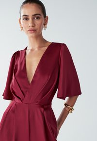 Mujer vistiendo un vestido de satén rojo profundo con escote en V y mangas acampanadas, accessorized con pendientes de aro dorados y pulseras gruesas.