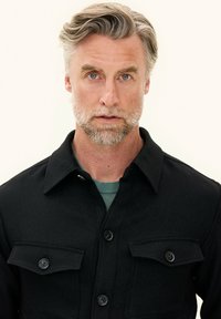 Black button-up jacket with two chest pockets, textured fabric, and a collar, worn over a green crew-neck sweater.