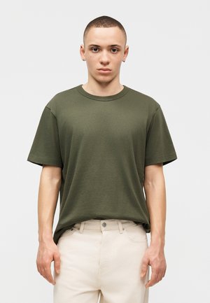 Νεαρός άνδρας με κοντά μαλλιά, που φοράει ελιάς πράσινο μπλουζάκι και εκρού παντελόνι, αντικρίζει ευθεία με ουδέτερη έκφραση σε απλό φόντο.