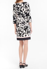 Robe à manches courtes en crème avec un motif floral noir, présente une coupe droite et un ourlet avec une bordure noire contrastante. Sandales à talons carrés noires.