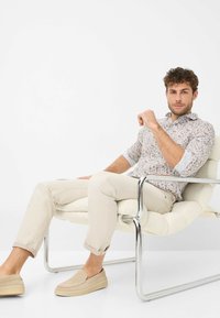 Bluse mit Blumenmuster, langärmlig, dazu hellbeige Hose und beigen Slipper, sitzend auf einem modernen Chaiselongue aus Chrom.