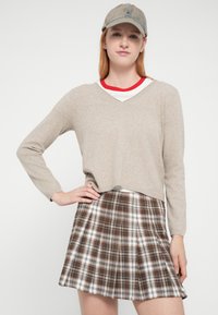 Pull en maille beige à col en V, superposé sur une chemise rayée rouge et blanche, associé à une jupe plissée à carreaux marron et une casquette délavée.