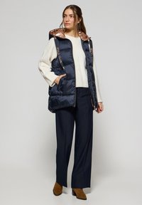 Gilet matelassé bleu marine avec une capuche brillante accentuée en cuivre, doté d'une fermeture éclair sur le devant, de poches latérales et du logo "POLO CLUB" le long des épaules.