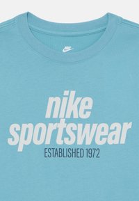 Šviesiai mėlynas medvilninis marškinėlis su ryškiais baltais užrašais „nike sportswear“ ir „ĮSTEIGTA 1972“ moderniu šriftu. Sukamo kaklo dizainas.