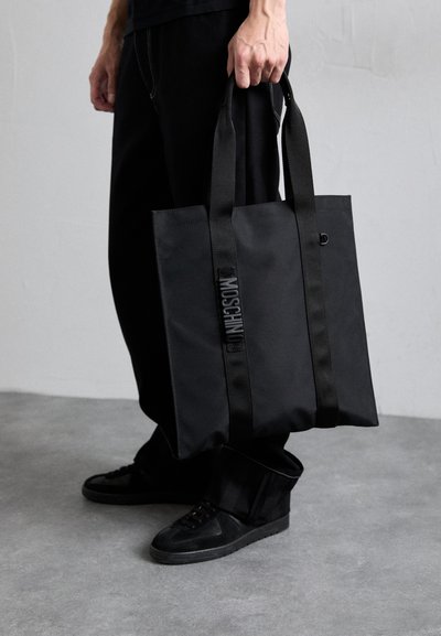 Black tote bag with a rectangular shape, made of durable fabric, featuring two sturdy straps and a logo patch along the center.