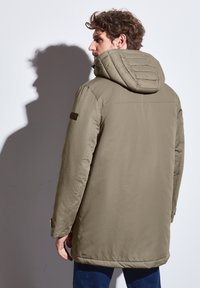 Khaki parka-jacka med en texturerad, vadderad huva. Har dragkedja, sidofickor och en texturerad axelpatch på vänster ärm.