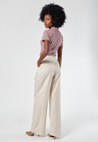 Camicia a maniche corte stampata con un motivo geometrico, abbinata a pantaloni beige a gamba larga con vita alta. Le calzature presentano una punta appuntita.