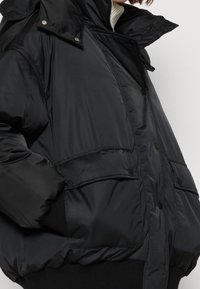 Chaqueta de plumas negra con acabado brillante, cuello alto y dos bolsillos frontales asegurados con botones a presión. Dobladillo de punto acanalado.