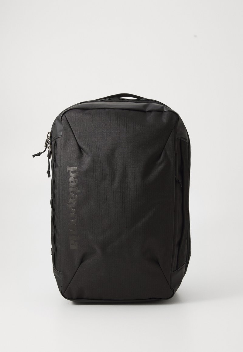 Mochila preta com um tecido texturizado, apresentando um fecho com zíper, bolsos laterais e um logótipo impresso na frente. Design compacto e retangular.
