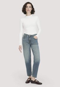 Weißes Langarmoberteil, hoch taillierte hellblaue Jeans mit geradem Bein und dunklen Schuhen. Das Model steht mit einer Hand in der Jeans-Tasche.