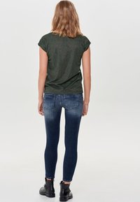 Mörkgrön, kortärmad topp med en subtil glans, kombinerad med mid-rise, skinny, blekta blå jeans. Modellen har på sig svarta ankelboots.
