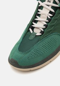 Sapatilhas verdes com uma mistura de malha texturizada e materiais de camurça. Projetadas com atacadores brancos, detalhes perfurados e uma sola de borracha preta.